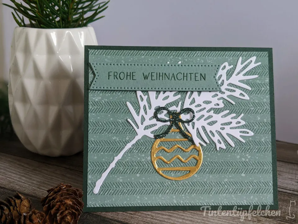 Weihnachtskarte mit gestempeltem grüner Hintergrund und weißem Folienschnitt von Tannenzweigen. Davor liegt ein goldener Stempelballon mit Wellenlinien, darüber ein schwarzer Glitterbogen. Oben ist ein gestickter Bandstreifen mit den Worten „FROHE WEIHNACHTEN“ angebracht. Im Hintergrund steht ein weißer geometrischer Topf mit einer kleinen Tanne und zwei Kiefernnadeln auf Holz.