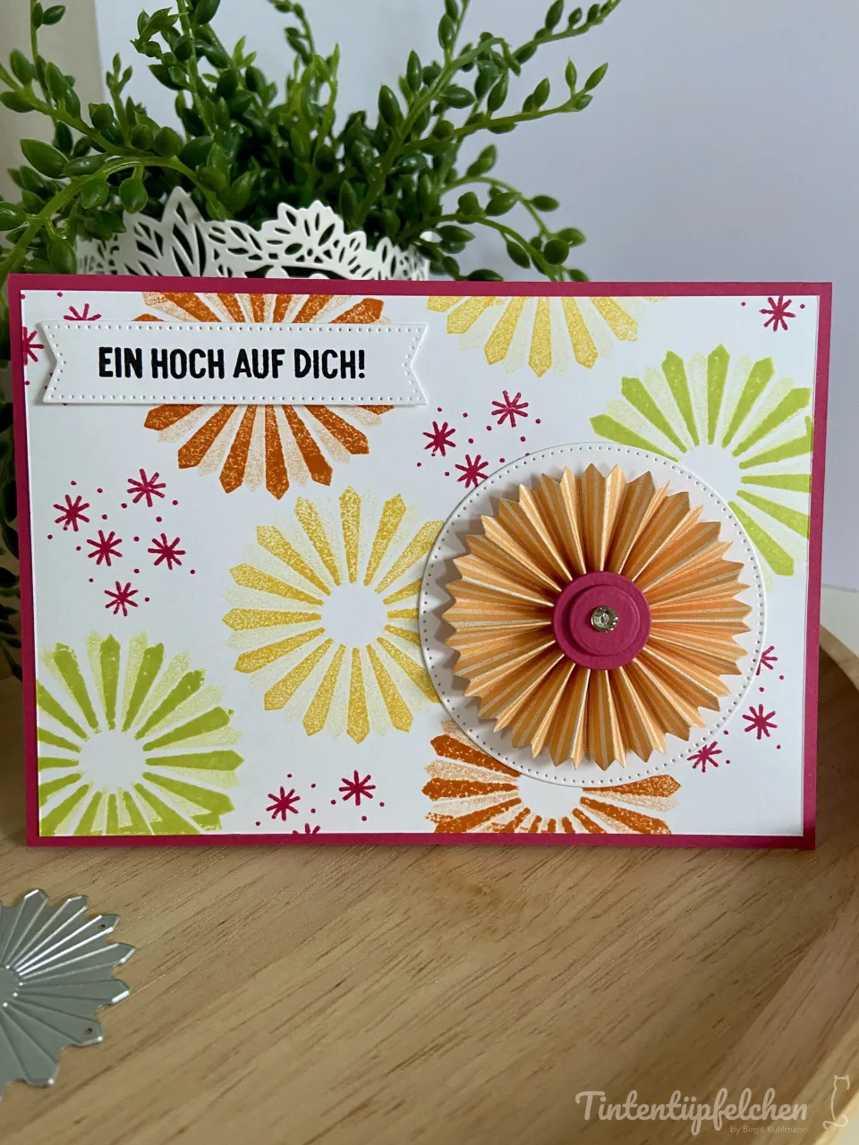 Eine selbstgebastelte Hochzeitskarte mit weißem Karton und rotem Rahmen, verziert mit gestempelten Sonnenblumen in Gelb, Orange und Grün sowie kleinen roten Sternen. Im Zentrum befindet sich eine dreidimensionale Papierblume aus orangefarbenem Pergament mit rosa Mittelpunkt und einem kleinen Glanzstein. Oben ist ein weißer Streifen mit dem Text „EIN HOCH AUF DICH!“ angebracht. Im Hintergrund steht eine grüne Pflanze in einer weißen Schablone, unten links liegt ein silbernes Stempelteil