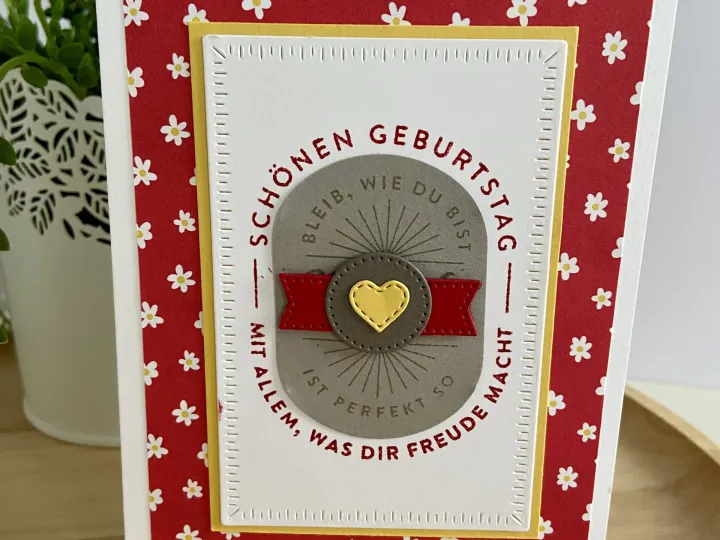 Eine gebastelte Geburtstagskarte mit rotem Hintergrundpapier, das mit kleinen weißen Blumen bedruckt ist. Im Zentrum befindet sich ein gestanzter Kreis in Grau mit einem gelben Herzen und einer roten Schleife, umgeben von der Aufschrift „Schönen Geburtstag – Bleib, wie du bist – Mit allem, was dir Freude macht“. Der Text ist in roter Farbe auf dem grauen Grund angeordnet und wird von einem weißen Kartonrahmen mit gestanztem Rand eingefasst.