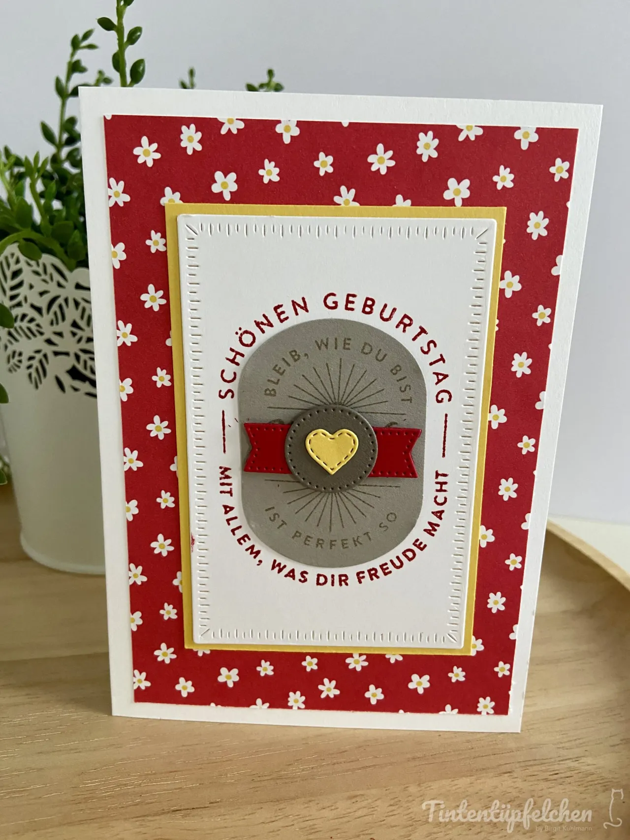 Eine gebastelte Geburtstagskarte mit rotem Hintergrundpapier, das mit kleinen weißen Blumen bedruckt ist. Im Zentrum befindet sich ein gestanzter Kreis in Grau mit einem gelben Herzen und einer roten Schleife, umgeben von der Aufschrift „Schönen Geburtstag – Bleib, wie du bist – Mit allem, was dir Freude macht“. Der Text ist in roter Farbe auf dem grauen Grund angeordnet und wird von einem weißen Kartonrahmen mit gestanztem Rand eingefasst.