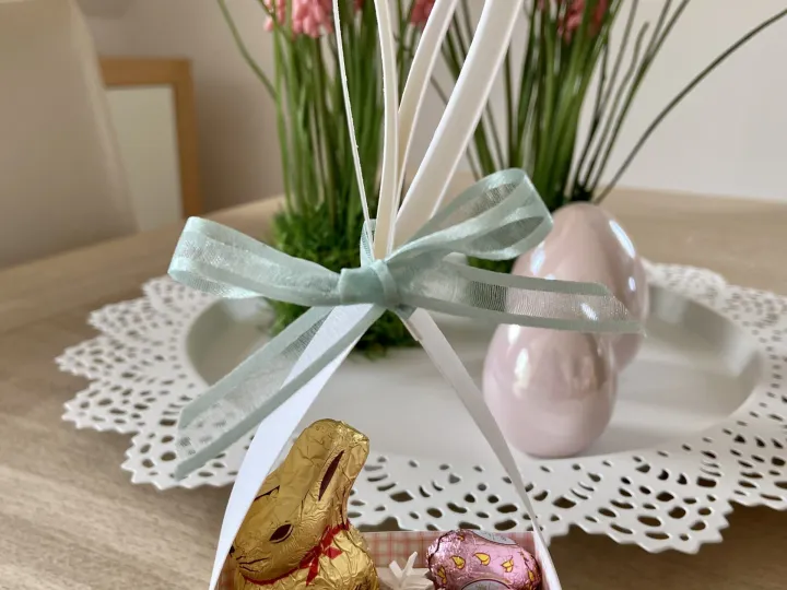 Ein selbstgebasteltes Oster-Eierkorb aus Papier mit grün-weißem Rastermuster und einem Holzherz als Verschluss, in dem ein Lindt-Schokobunny und zwei rosa Schokoeier liegen. Weiße Papierohren sind oben befestigt und von einer hellgrünen Seidenbandkrawatte umschlossen. Im Hintergrund steht eine Kerzenblume mit pinken Blüten auf einem weißen Teller über einem Holztisch.