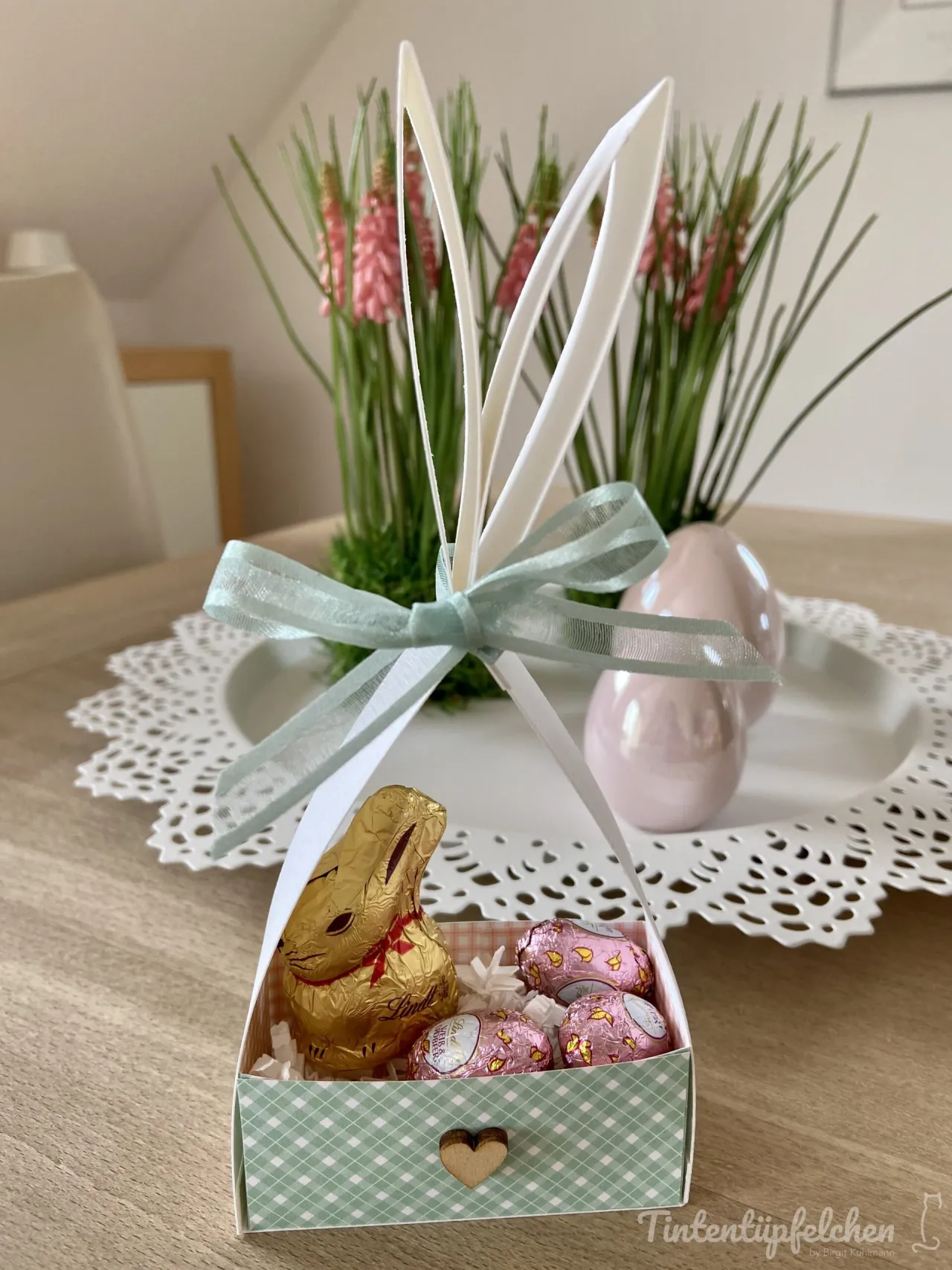 Ein selbstgebasteltes Oster-Eierkorb aus Papier mit grün-weißem Rastermuster und einem Holzherz als Verschluss, in dem ein Lindt-Schokobunny und zwei rosa Schokoeier liegen. Weiße Papierohren sind oben befestigt und von einer hellgrünen Seidenbandkrawatte umschlossen. Im Hintergrund steht eine Kerzenblume mit pinken Blüten auf einem weißen Teller über einem Holztisch.