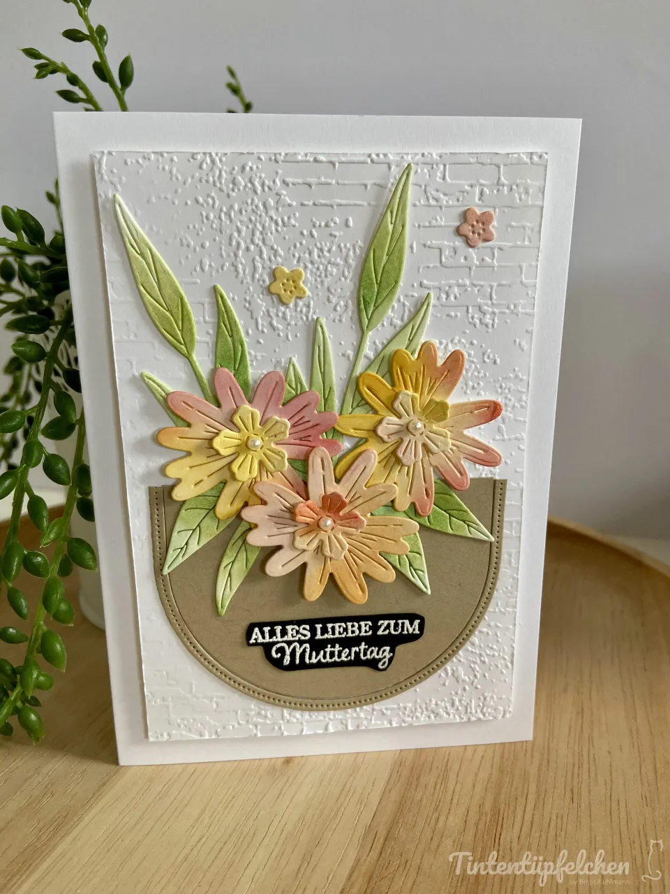 Eine gestempelte Muttertagskarte mit weißem Hintergrund und Bruchstein-Textur. Drei dreidimensionale Blumen in Pastellfarben (Lila, Gelb, Rosa) mit Perlen als Blütenmitte sind in einer braunen Halbkreisform angeordnet, umgeben von grünen Blättern. Unten steht der schwarze Text „Alles Liebe zum Muttertag". Links im Bild ist eine Grüne Pflanze sichtbar.