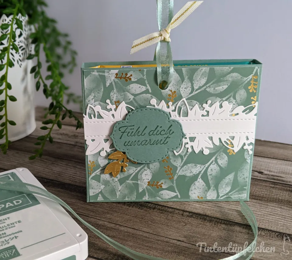 Ein selbstgebastelter Geschenkbeutel mit grünem Blattmuster und goldenen Akzenten, verziert mit weißem gestochenem Papierstreifen und einem ovalen Aufkleber mit der Aufschrift „Fühl dich umarmt". Ein Seidenband ist oben gebunden, unten liegt ein Tintenpad auf Holz.