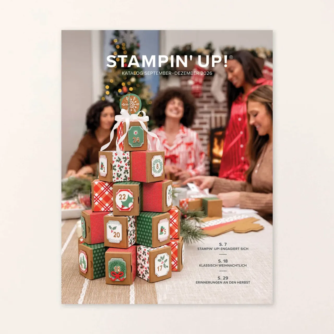 Ein Stempelkatalog für September bis Dezember 2026 zeigt eine Pyramide aus handgestempelten Adventskalender-Boxen in Rot, Grün und Braun mit Zahlen auf Etiketten. Im Hintergrund sitzen vier Frauen bei einem Tisch mit Bastelmaterial; im unscharfen Hintergrund ist ein geschmückter Weihnachtsbaum zu erkennen.