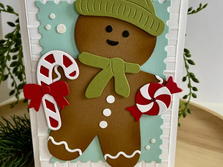 Eine handgemachte Weihnachtskarte mit einem gestempelten und farbig bemalten Gingerbread-Man in brauner Farbe, der eine grüne Mütze und ein Gewand trägt. Der Mann hält einen roten Zuckerstangen-Stempel mit weißem Streifen und ein rotes Zuckerpapier-Spiral-Motiv. Im Hintergrund ist ein hellblauer gestellter Bereich mit weißen Punkten zu sehen, umgeben von einem weißen Rastermuster auf der Karte. Kleine rote und weiße Zuckerpapier-Elemente liegen vor der Karte auf dem Holztisch.