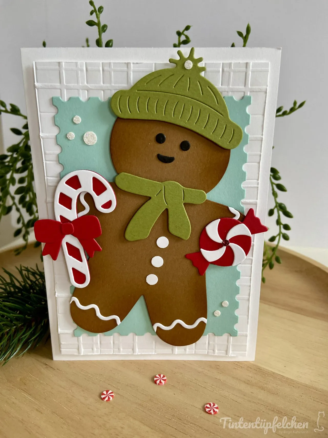 Eine handgemachte Weihnachtskarte mit einem gestempelten und farbig bemalten Gingerbread-Man in brauner Farbe, der eine grüne Mütze und ein Gewand trägt. Der Mann hält einen roten Zuckerstangen-Stempel mit weißem Streifen und ein rotes Zuckerpapier-Spiral-Motiv. Im Hintergrund ist ein hellblauer gestellter Bereich mit weißen Punkten zu sehen, umgeben von einem weißen Rastermuster auf der Karte. Kleine rote und weiße Zuckerpapier-Elemente liegen vor der Karte auf dem Holztisch.