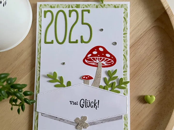 Eine selbstgebastelte Karte mit dem Text „2025“ in grüner Schrift und einem roten Pilz auf weißem Papier, umgeben von grünen Blättern und kleinen Diamanten. Unten ist ein gestickter Umschlag mit der Aufschrift „Viel Glück!“, verziert mit einer silbernen Schleife und einem glitzernden Vierblütigerkraut.