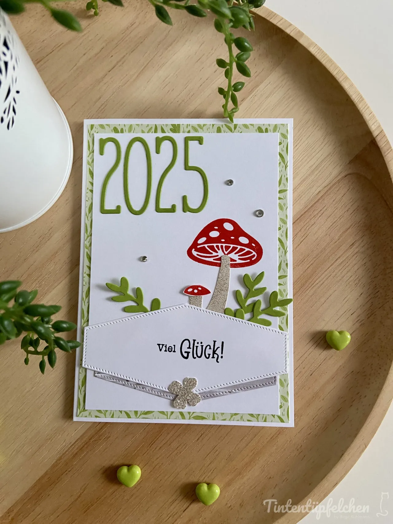 Eine selbstgebastelte Karte mit dem Text „2025“ in grüner Schrift und einem roten Pilz auf weißem Papier, umgeben von grünen Blättern und kleinen Diamanten. Unten ist ein gestickter Umschlag mit der Aufschrift „Viel Glück!“, verziert mit einer silbernen Schleife und einem glitzernden Vierblütigerkraut.