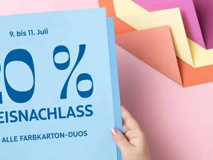 Eine Person hält ein blaues Flyer mit der Aufschrift „20 % PREISNACHLASS AUF ALLE FARBKARTON-DUOS“ für die Zeit vom 9. bis 11. Juli. Im Hintergrund liegen gestapelte farbige Papierkarten in Rosa, Lila, Gelb und Orange auf einem hellrosa Untergrund.