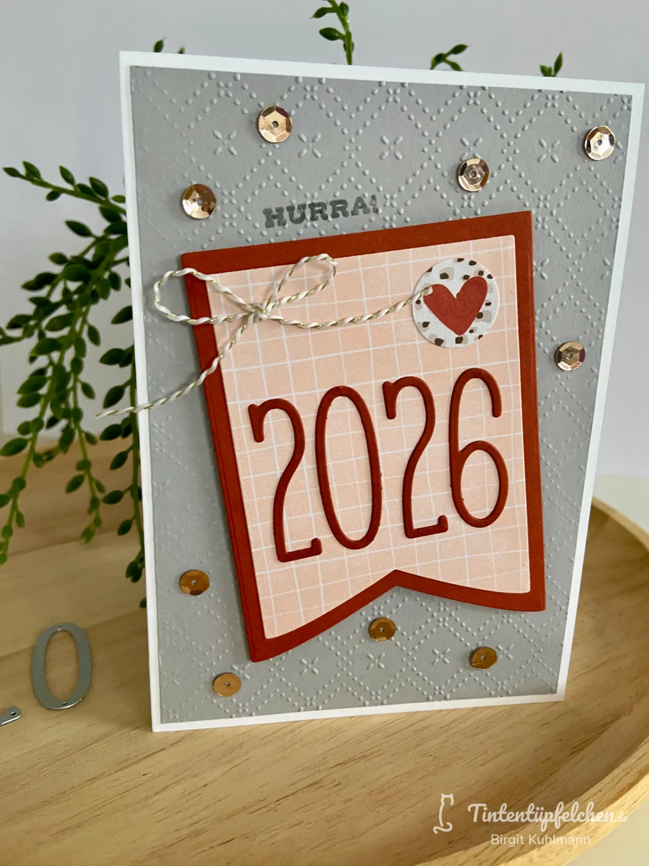 Eine gebastelte Grußkarte mit gestempeltem grauem Hintergrund und goldenen Glitzern, die ein orangefarbenes Etikett mit der Jahreszahl 2026 und einem roten Herzen zeigt. Ein weißer Schnurknoten ist oben links befestigt.
