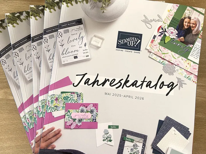 Ein Stapel Stampin' Up Jahreskataloge Mai 2025 bis April 2026 liegt auf einem Holztisch, umgeben von Bastelmaterialien wie Stempeln, Farbtinten und floralen Papierelementen. Ein weißes Doilieschneidepapier ist oben sichtbar, während unten eine Person die Kataloge hält.