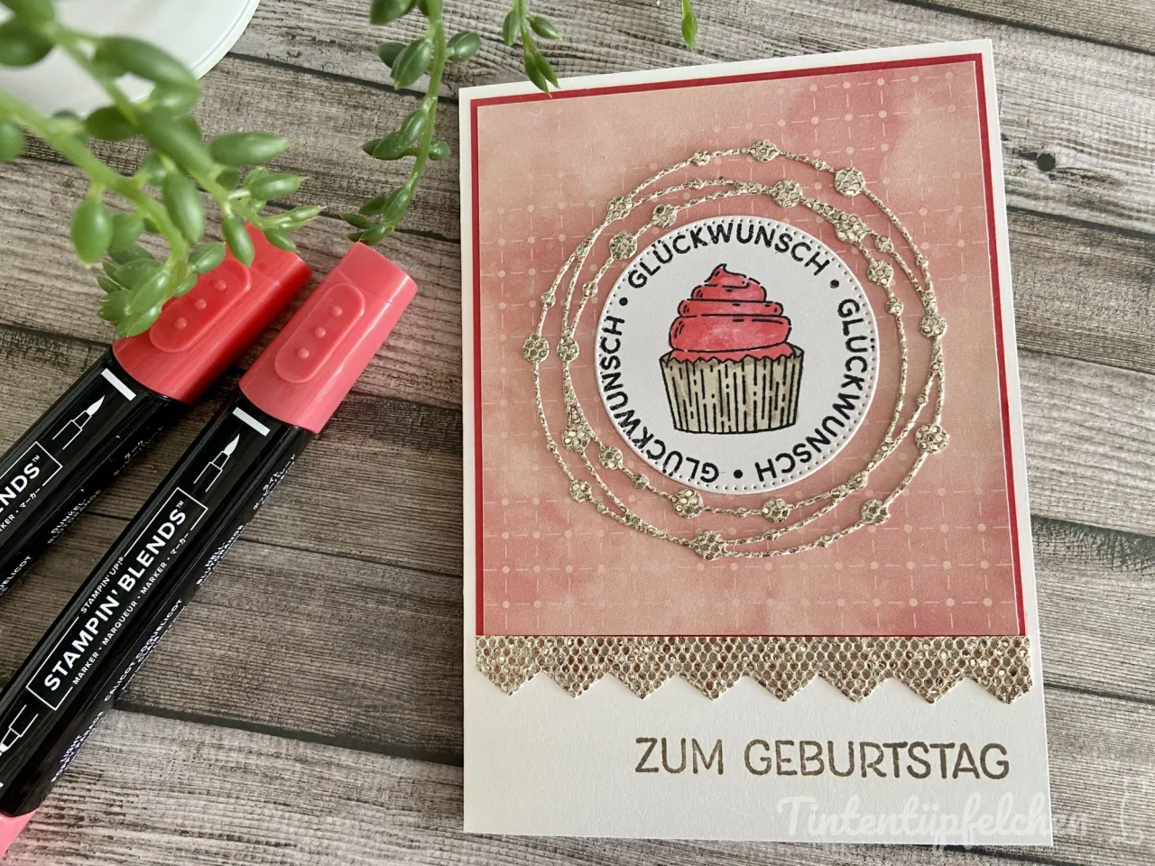 Eine Geburtstagskarte mit einem gestempelten Cupcake in Rosa auf weißem Kreis, umgeben von silbernen Sticker-Verzierungen und dem Wort „GLÜCKWUNSCH“ im Kreis. Der Hintergrund der Karte ist rosa mit feinem Rastermuster, unten befindet sich ein goldener Spitzenrand und die Großbuchstaben „ZUM GEBURTSTAG“. Links liegen zwei rote Stampin’ Blends Marker auf Holzoptik-Hintergrund mit grüner Pflanze oben links.