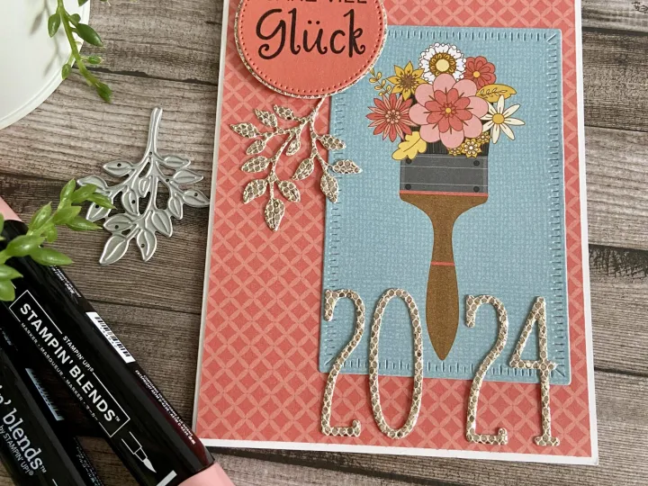 Eine handbastelte Grußkarte mit einem gestempelten Bild eines Pinsels, der eine Blumenvase hält und die Zahl 2024 darstellt. Der Hintergrund ist in Rosa mit geometrischem Muster gehalten, oben befindet sich ein abgerundeter Aufkleber mit der Aufschrift „Ganz viel Glück“. Umgebend sind Stempelmarkierer und ein Metallstempel sichtbar auf einem Holzoberflächenhintergrund.