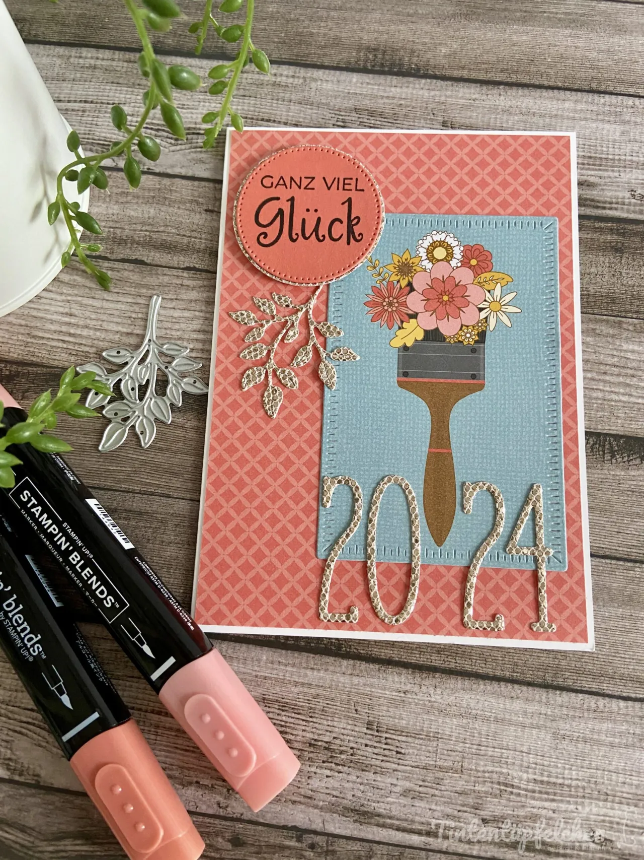 Eine handbastelte Grußkarte mit einem gestempelten Bild eines Pinsels, der eine Blumenvase hält und die Zahl 2024 darstellt. Der Hintergrund ist in Rosa mit geometrischem Muster gehalten, oben befindet sich ein abgerundeter Aufkleber mit der Aufschrift „Ganz viel Glück“. Umgebend sind Stempelmarkierer und ein Metallstempel sichtbar auf einem Holzoberflächenhintergrund.