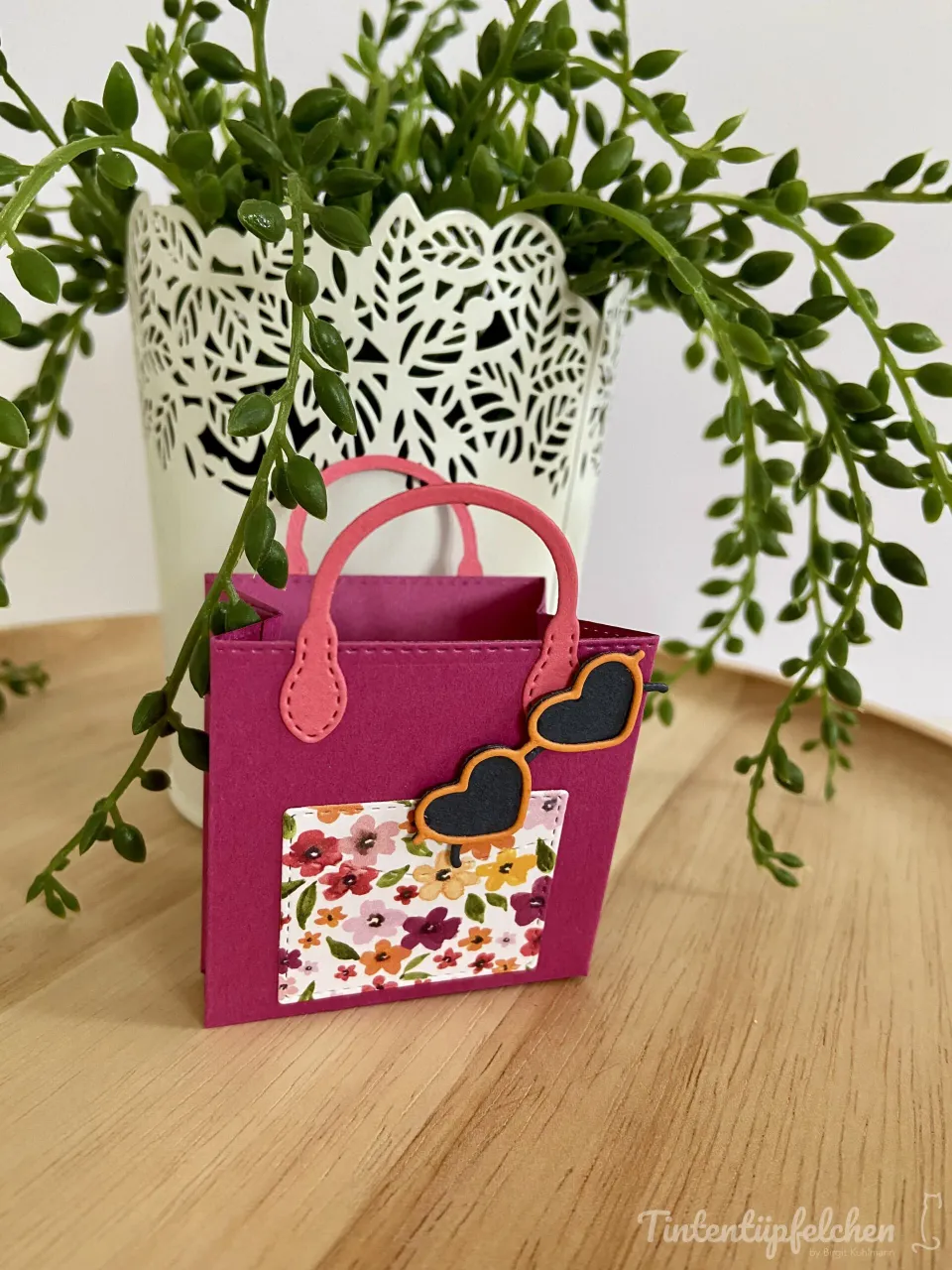 Ein kleines, rosa Basteltasch mit runden Trägern und gestickten Nähten steht auf einem Holltisch. Auf der Vorderseite ist ein floraler Ausschnitt angebracht, über dem eine Herzform in Schwarz mit orangefarbenem Rahmen befestigt ist. Im Hintergrund befindet sich eine Pflanze in einer weißen Schere aus Papier.