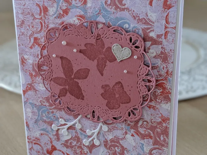 Eine Grußkarte mit einem gemusterten Hintergrund in Rosa-, Rot- und Lilatönen. Darauf liegt eine rosafarbene, gestanzte Kreisform mit Blumenmotiven und kleinen Perlen. Ein kleiner Herzanhänger und ein Zweig mit Blättern sind ebenfalls angebracht. Unten rechts befindet sich ein kleiner, rechteckiger Aufleger mit dem Spruch "An dich gedacht, für dich gemacht!".