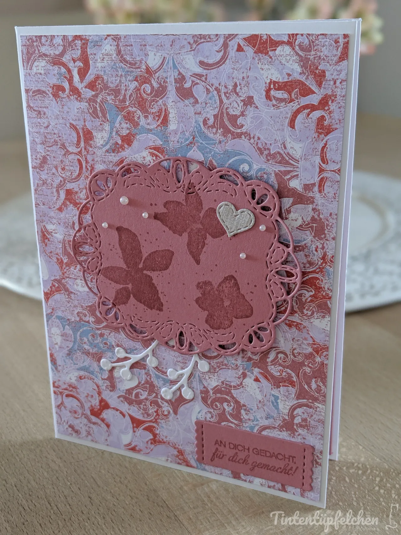 Eine Grußkarte mit einem gemusterten Hintergrund in Rosa-, Rot- und Lilatönen. Darauf liegt eine rosafarbene, gestanzte Kreisform mit Blumenmotiven und kleinen Perlen. Ein kleiner Herzanhänger und ein Zweig mit Blättern sind ebenfalls angebracht. Unten rechts befindet sich ein kleiner, rechteckiger Aufleger mit dem Spruch "An dich gedacht, für dich gemacht!".