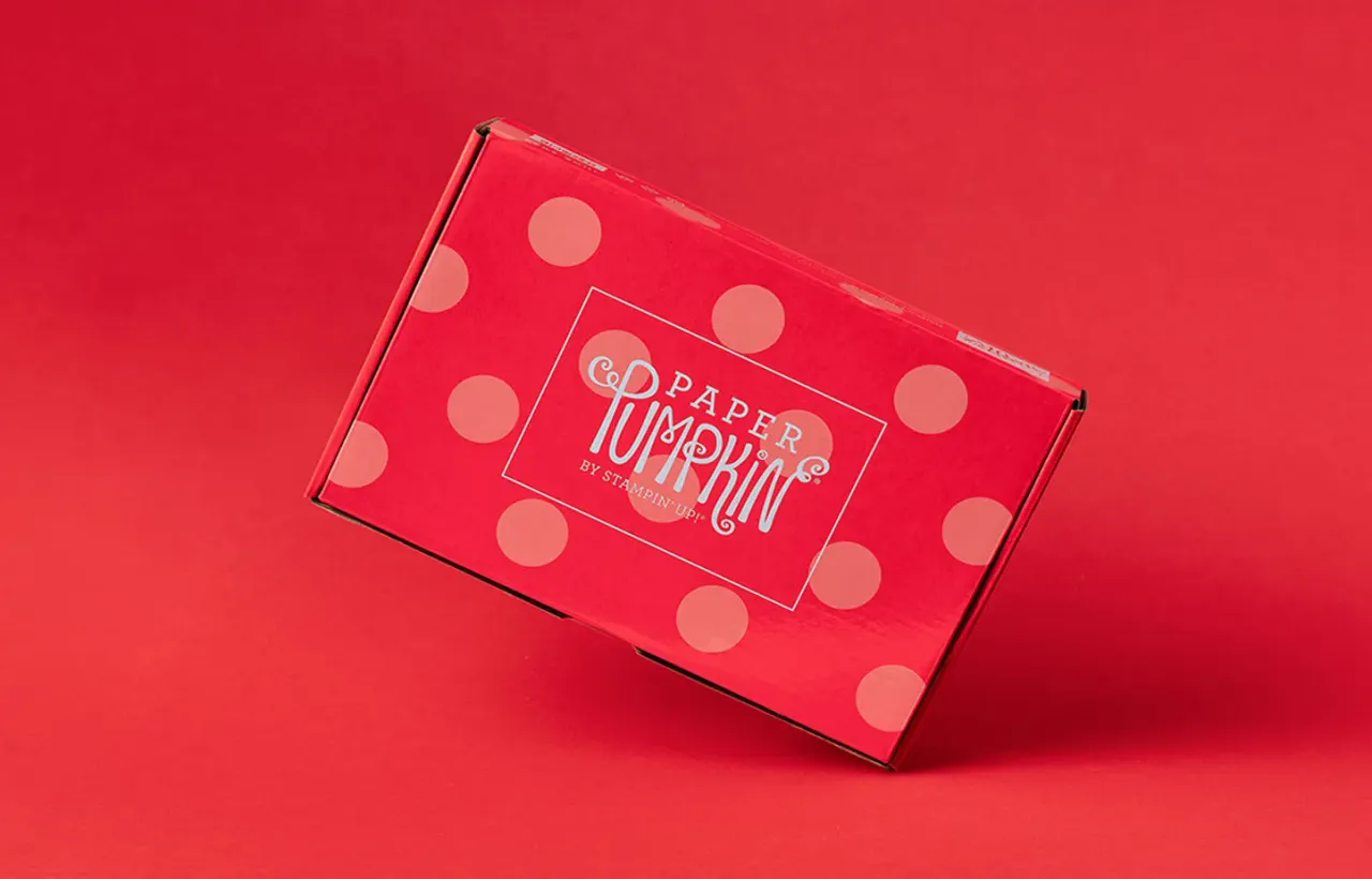 Ein roter Karton mit hellrosa Polka-Dots, auf dem ein weißer Rahmen den Text „PAPER PUMPKIN BY STAMPIN UP“ in verspielten Schriften zeigt. Das Bild ist schräg aufgenommen und wirft einen leichten Schatten auf den roten Hintergrund.