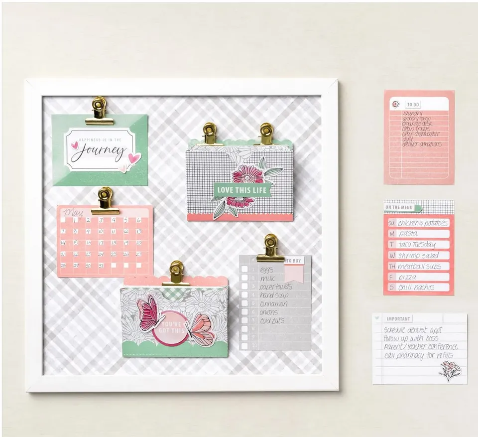 Ein weißer Rahmen mit einem gestreiften Hintergrund hält verschiedene Papiersteckkarten in goldenen Klammern. Darin sind ein rosa Kalender, eine Karte mit Schmetterlingen und Blumen sowie Listen für Einkaufs- und To-Do-Listen zu sehen. Neben dem Rahmen hängen weitere kleine Notizen an der Wand.