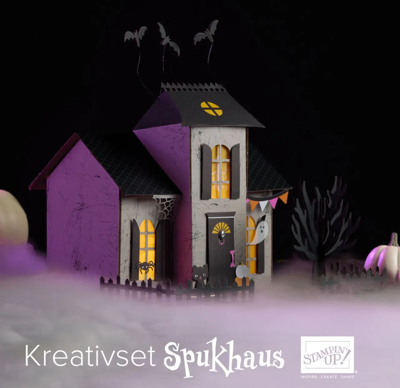 Ein aus Papier und Stempeln gefertigtes Halloween-Spukhaus mit lila und grauen Wänden, gelben Fenstern, schwarzen Dächern und fliegenden Fledermäusen auf dem Dach. Um das Haus herum stehen kleine grüne Bäume, ein weißer Geist an der Wand und eine Spinnweben-Dekoration; im Vordergrund liegt weißes Papier als Nebel.