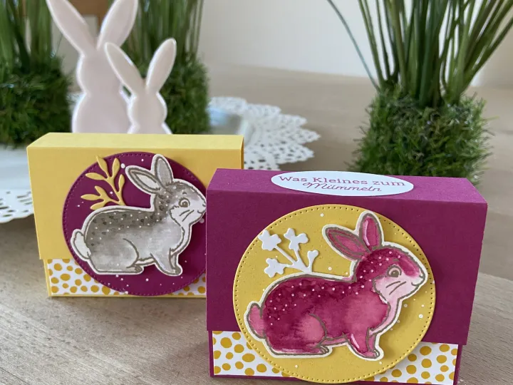Zwei kleine Geschenkkartenboxen mit bunten Hasen-Motiven aus gestempeltem Papier stehen auf einem Holttisch. Die Box im Vordergrund ist in lila und gelb gehalten, zeigt einen rosa Hasen auf gelbem Kreisgrund mit weißem Stempelwerk und trägt oben eine weiße Aufkleber-Textur mit der Aufschrift „Was Kleines zum Mümmeln“. Im Hintergrund steht eine weitere Box in Gelb mit einem grauen Hasenbild auf lila Grund, umgeben von grünen Pflanzen und weißen Hase-Figuren.