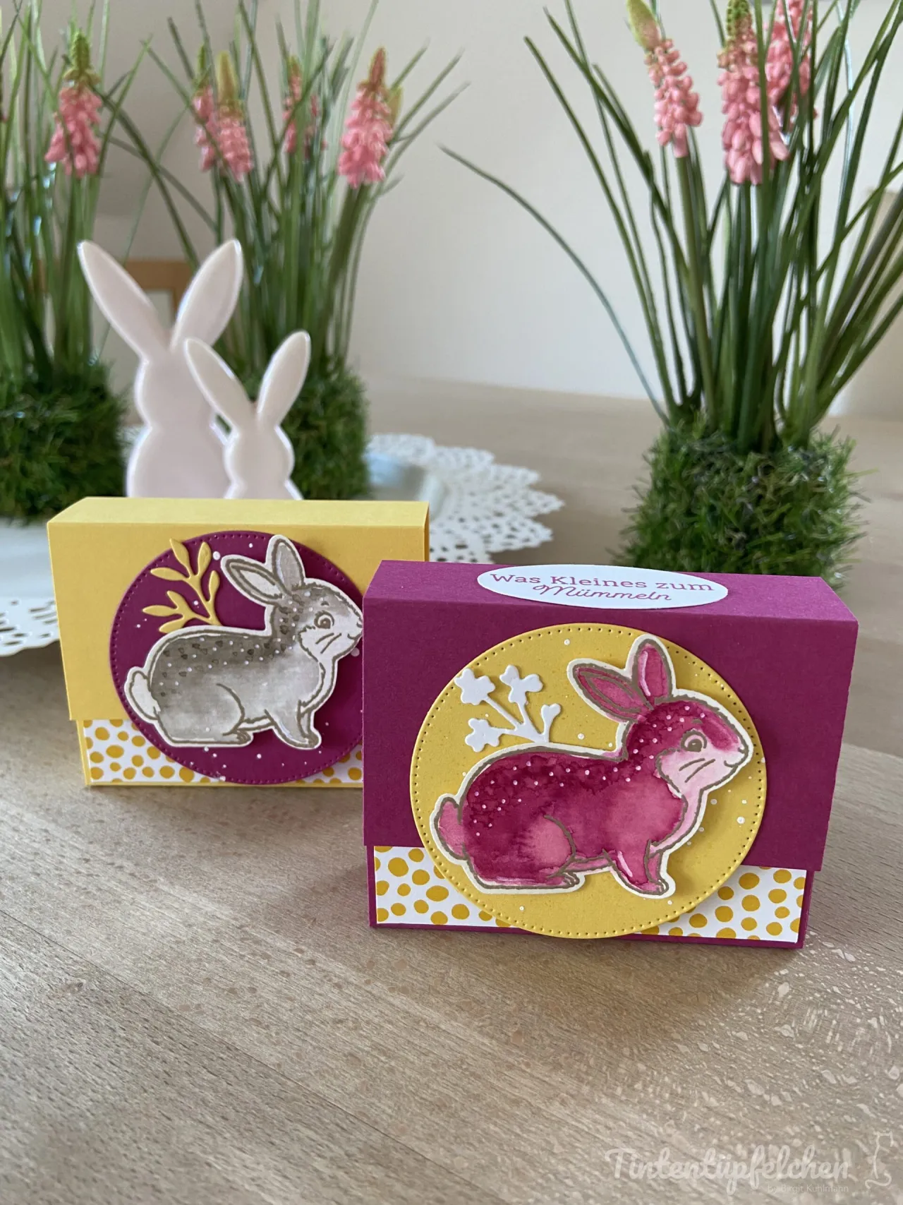 Zwei kleine Geschenkkartenboxen mit bunten Hasen-Motiven aus gestempeltem Papier stehen auf einem Holttisch. Die Box im Vordergrund ist in lila und gelb gehalten, zeigt einen rosa Hasen auf gelbem Kreisgrund mit weißem Stempelwerk und trägt oben eine weiße Aufkleber-Textur mit der Aufschrift „Was Kleines zum Mümmeln“. Im Hintergrund steht eine weitere Box in Gelb mit einem grauen Hasenbild auf lila Grund, umgeben von grünen Pflanzen und weißen Hase-Figuren.