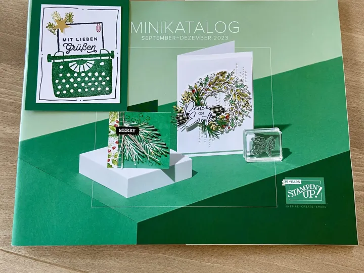 Ein offener Minikatalog von Stampin' Up! mit dem Titel „September–Dezember 2023“ zeigt vier Bastelprojekte in grünen und weißen Tönen. Darunter sind eine Karte mit einer gestempelten Schreibmaschine, eine weitere mit einem Tannenzweig-Motiv, eine Weihnachtskranzkarte mit der Aufschrift „Peace on Earth“ sowie ein transparenter Stempelblock mit Blattmotiven. Oben ist ein weißer Papiereinkuss sichtbar, unten befindet sich das Logo von Tintentüpfelchen.