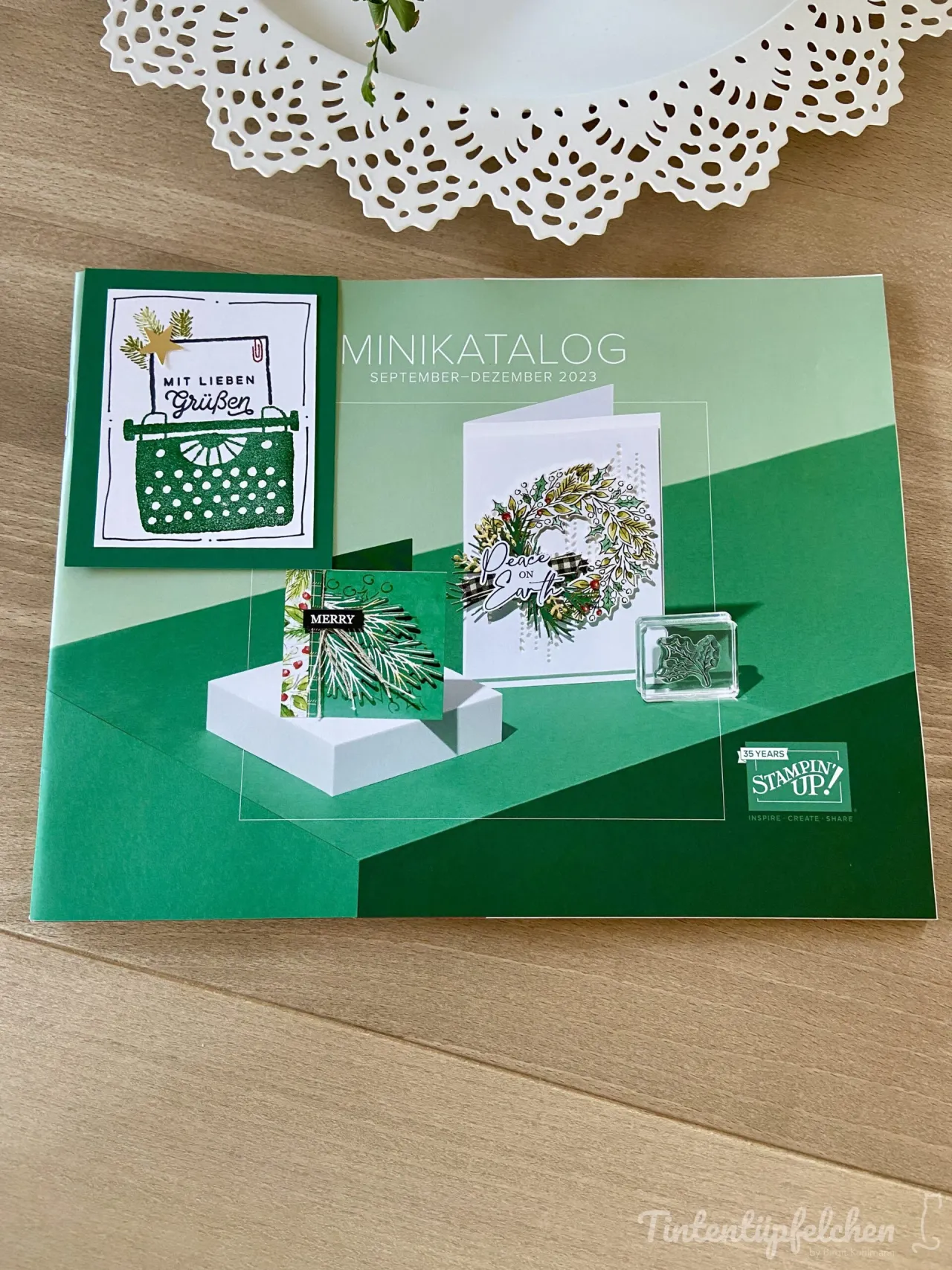 Ein offener Minikatalog von Stampin' Up! mit dem Titel „September–Dezember 2023“ zeigt vier Bastelprojekte in grünen und weißen Tönen. Darunter sind eine Karte mit einer gestempelten Schreibmaschine, eine weitere mit einem Tannenzweig-Motiv, eine Weihnachtskranzkarte mit der Aufschrift „Peace on Earth“ sowie ein transparenter Stempelblock mit Blattmotiven. Oben ist ein weißer Papiereinkuss sichtbar, unten befindet sich das Logo von Tintentüpfelchen.