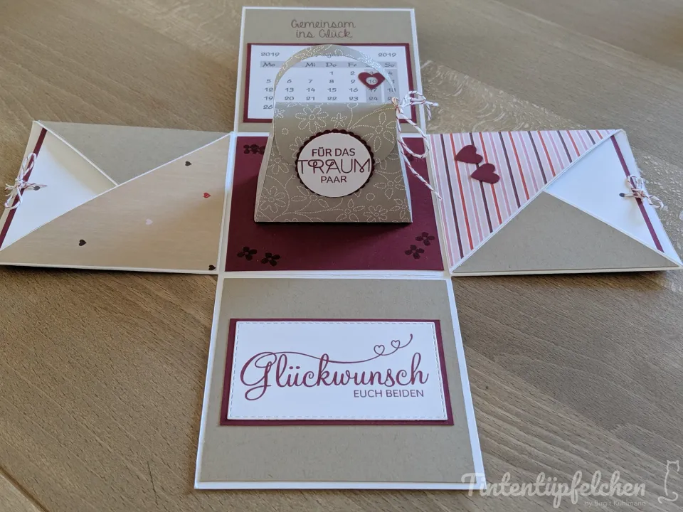 Eine aufklappbare Box in Beige und Bordeauxrot mit einem Kalenderblatt, einer kleinen Geschenktüte und der Aufschrift "Glückwunsch euch beiden".