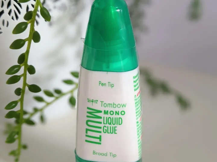 Ein grüner Tombow Mono Liquid Glue-Stift mit breiter Spitze steht auf weißem Untergrund. Im Hintergrund ist eine unscharfe grüne Pflanze und ein weißes Blatt aus Papier zu erkennen. Der Stift hat einen transparenten grünen Behälter mit weißem Etikett, das die englischen Bezeichnungen „Pen Tip“ und „Broad Tip“ sowie den Markennamen enthält.