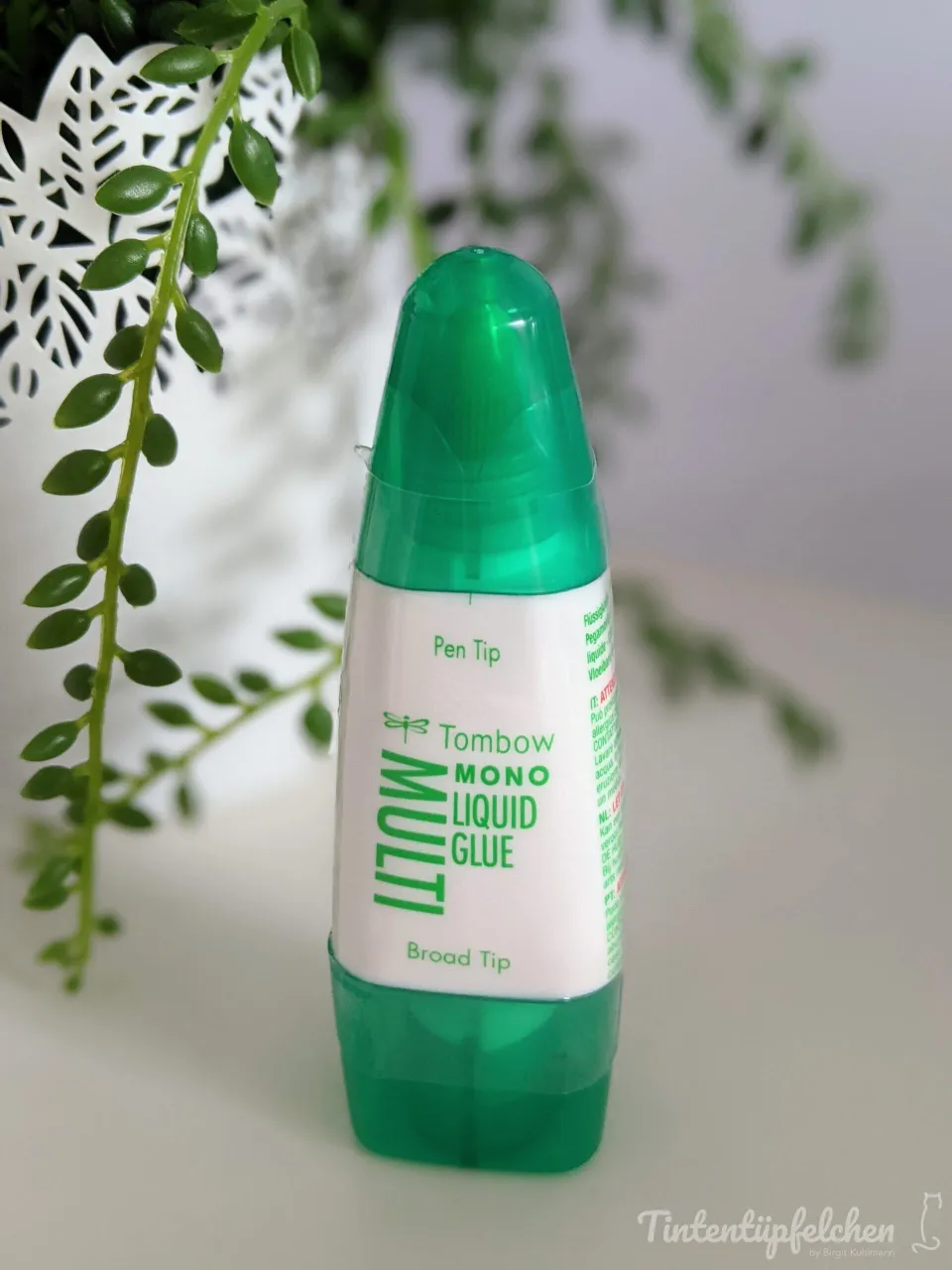 Ein grüner Tombow Mono Liquid Glue-Stift mit breiter Spitze steht auf weißem Untergrund. Im Hintergrund ist eine unscharfe grüne Pflanze und ein weißes Blatt aus Papier zu erkennen. Der Stift hat einen transparenten grünen Behälter mit weißem Etikett, das die englischen Bezeichnungen „Pen Tip“ und „Broad Tip“ sowie den Markennamen enthält.