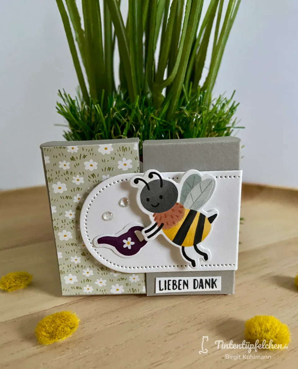 Ein kleines Bastelstück aus Pappe mit weißem und grauem Hintergrund, verziert mit einem gestempelten Bienenmotiv in Gelb, Schwarz und Grau, das eine kleine Pflanze gießt. Der Text „LIEBEN DANK“ ist auf einem grauen Streifen unten angebracht, umgeben von kleinen gelben Deko-Kugeln und einer grünen Pflanze im Hintergrund.