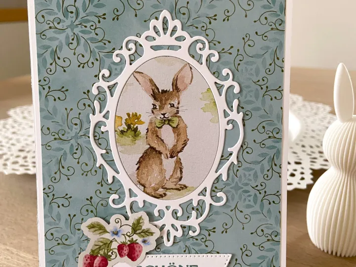 Eine Ostergeschenkkarte mit blau-grünem Blattmuster als Hintergrund und einem weißen filigranen Ovalrahmen, der ein gestochenes Bild eines Hasen mit grüner Schleife zeigt. Unten ist ein weißer Streifen mit dem Text „SCHÖNE OSTERN“ angebracht, daneben eine Aufkleber-Abbildung von Erdbeeren und kleinen Blumen.
