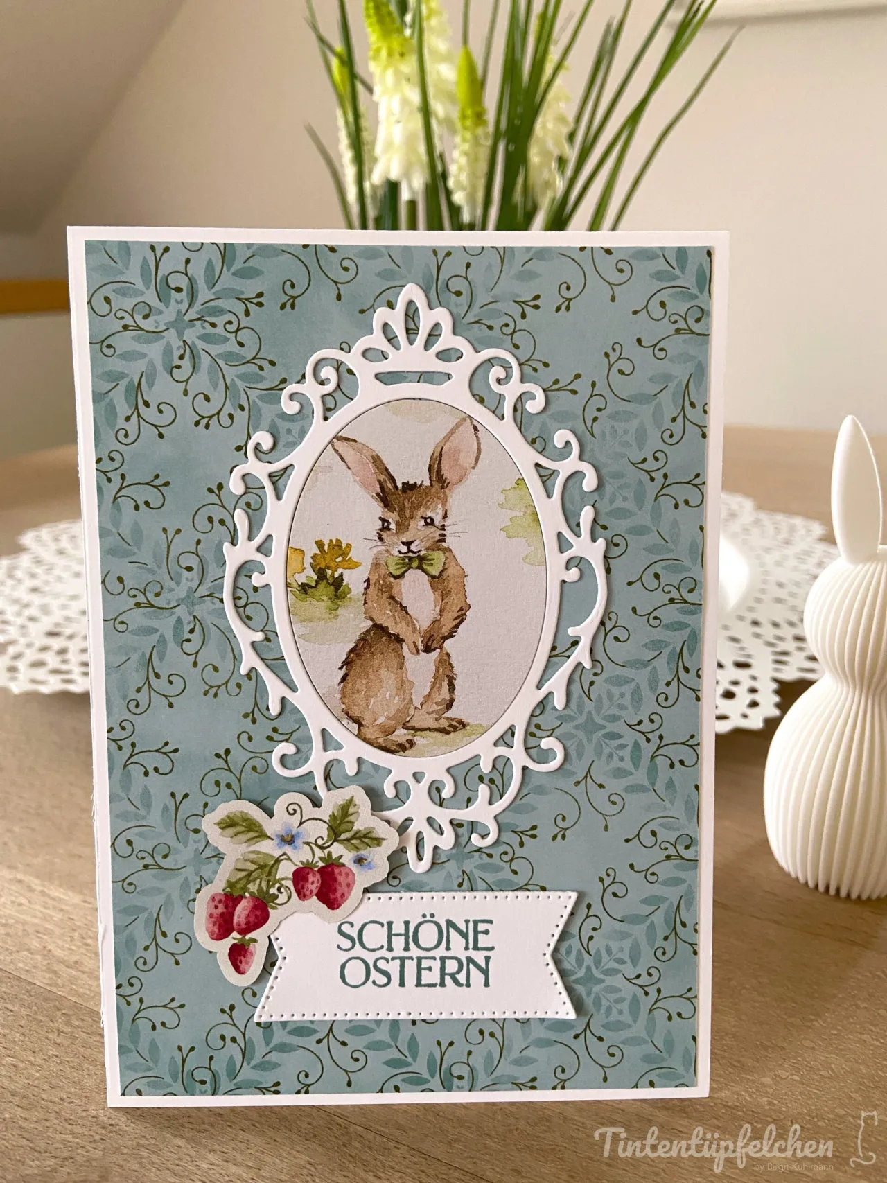 Eine Ostergeschenkkarte mit blau-grünem Blattmuster als Hintergrund und einem weißen filigranen Ovalrahmen, der ein gestochenes Bild eines Hasen mit grüner Schleife zeigt. Unten ist ein weißer Streifen mit dem Text „SCHÖNE OSTERN“ angebracht, daneben eine Aufkleber-Abbildung von Erdbeeren und kleinen Blumen.