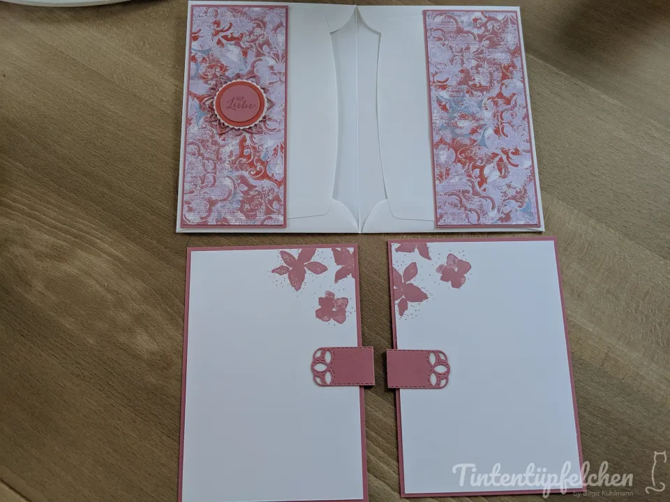 Eine aufklappbare Karte in Weiß mit zwei Einstecktaschen. Die Außenseiten sind mit einem gemusterten Papier in Rosa und Lila beklebt und mit einem roten Kreis mit der Aufschrift "Liebe" verziert. Daneben liegen zwei weiße Karten mit rosa Blumenstempelmotiven und einer kleinen rosa Schlaufe.