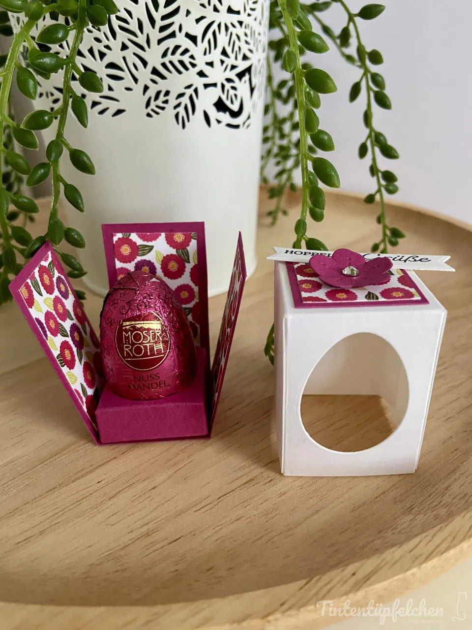Ein weißer Karton mit einem ovalen Ausschnitt dient als Geschenkbox und ist mit rosa Blumenschildern verziert. Eine kleine Karte mit der Aufschrift „Hoppe Grüß" trägt eine rosa Blütenform mit Glanzstein in der Mitte. Daneben liegt ein aufklappbarer Karton, der eine rosafarbene Schokolade enthüllt, die mit floralen Mustern bedruckt ist.