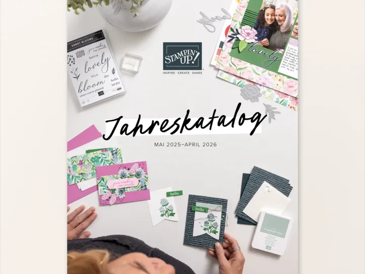 Auf einem hellen Tisch liegen verschiedene Bastelmaterialien von Stampin' Up!, darunter Stempelsets mit floralen Motiven und Grußworten, sowie fertige Karten in Rosa, Grün und Blau. Eine Person hält eine Karte mit gestempelten Blumen und einer grünen Beschriftung „hello". Im Hintergrund sind weitere Papierstücke, ein kleiner Topf mit grünen Pflanzen und silberne Schriftzüge zu sehen. Der Text „Jahreskatalog MAI 2025–APRIL 2026" steht zentral auf dem Bild.