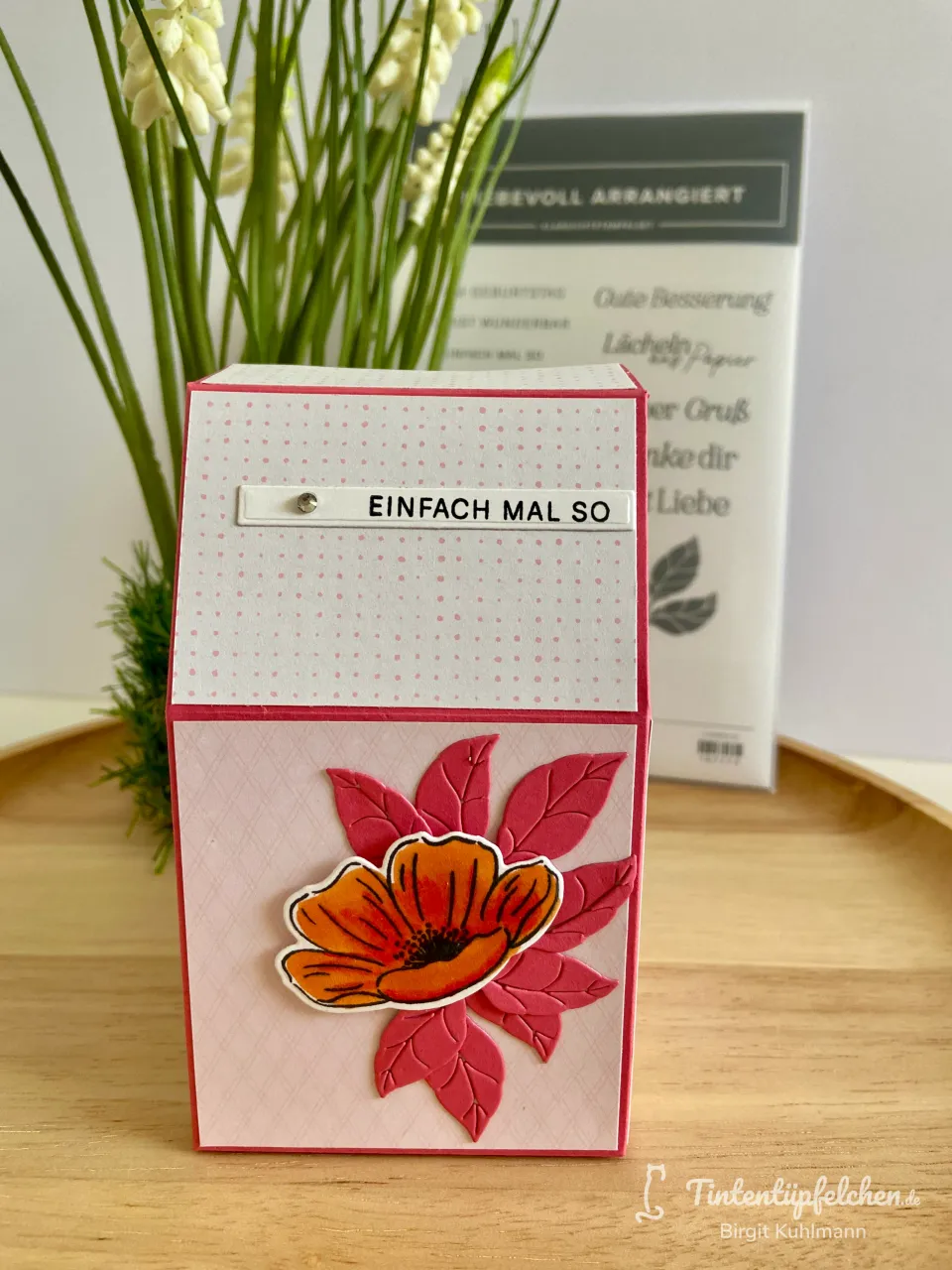 Ein selbstgebasteltes Kasten-Kartenstück mit weißem und pinkfarbenem Punktpapier sowie einem gestempelten, gefärbten Papierenblumenmotiv in Orange und Pink. Der Text „EINFACH MAL SO“ ist auf einer weißen Streifenetikette angebracht. Im Hintergrund steht ein Stempelset mit deutschen Grußworten neben grünen Pflanzenstängeln.