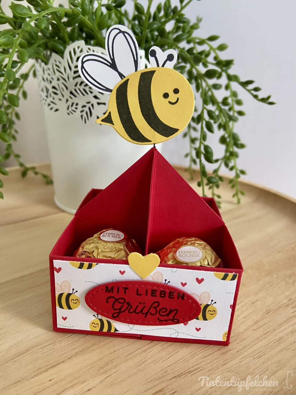 Ein aus rotem Karton gebasteltes Geschenkbox mit dreieckigem Dach, darin zwei Ferrero Rocher-Schokovliesen. Die Vorderseite ist mit weißem Papier bedruckt, das Bienen und rote Herzen zeigt; ein roter ovaler Aufkleber trägt die Aufschrift „MIT LIEBEN Grüßen“ in schwarzer Schrift, darüber liegt ein gelbes Herz. Oben ragt eine gestanzte Bienenfigur mit schwarz-weiß gefärbten Flügeln heraus. Im Hintergrund steht eine grüne Zimmerpflanze in einem