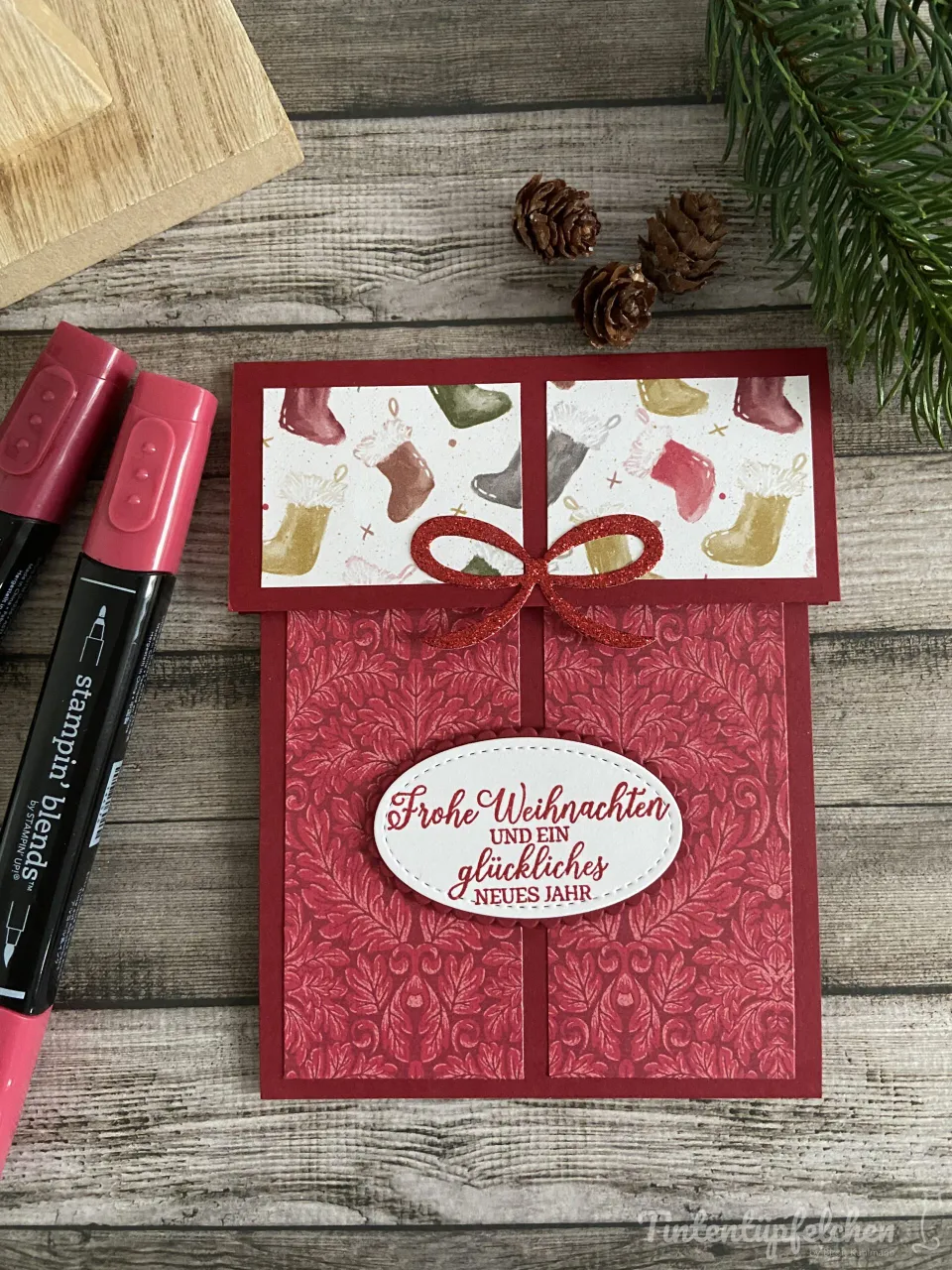 Eine handgestempelte Weihnachtskarte mit rotem Glanzpapier und einem gestochenen Muster, verziert mit zwei weißen Etiketten, die „Frohe Weihnachten und ein glückliches Neues Jahr“ tragen. Ein roter Glanzstift in Form eines Schleifens ist auf der Karte angebracht. Links liegen zwei schwarze Marker mit rosa Kapen, daneben befinden sich kleine Kiefernnadeln und eine Nadeln-Tanne auf einem Holzuntergrund.