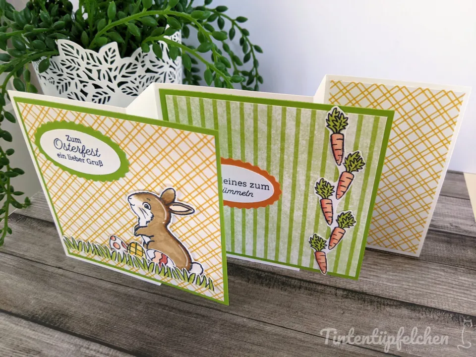 Drei Osterkarten mit gestempelten Motiven und handgefarbten Details stehen auf einem Holztisch. Die linke Karte zeigt einen braunen Hasen auf grünem Gras mit gelbem Eierschalen-Muster, der Text „Zum Osterfest ein lieber Gruß“ ist in einer grünen Spruchblase platziert. Die mittlere Karte hat vertikale Streifen in hellgrün und Weiß und trägt fünf orangefarbene Möhren mit grüner Krone; der Text „…eines zum Himmeln“ ist teilweise sichtbar. Rechts steht eine weitere Karte