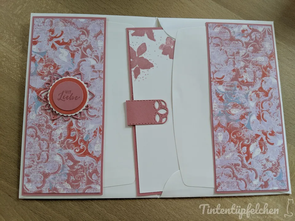 Eine weiße Klappkarte mit zwei schmalen, länglichen Seitenflügeln aus gemustertem Papier in Rosa, Rot und Blau. In der Mitte befindet sich ein schmaler Einsteckstreifen mit Blütenstempelungen in Rosa und einem kleinen Verschluss.