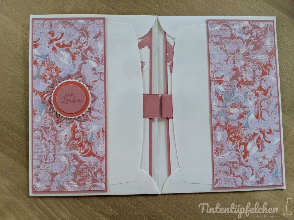 Eine gefaltete Karte mit rosa und lila gemusterten Papierstreifen, die durch eine zentrale Öffnung geschlossen werden. Ein runder Aufdruck mit der Aufschrift „Mit Liebe“ ist linksseitig angebracht. Die Innenseite zeigt weiße Flächen mit vertikalen rosa Streifen und einer kleinen rosa Schlinge in der Mitte.