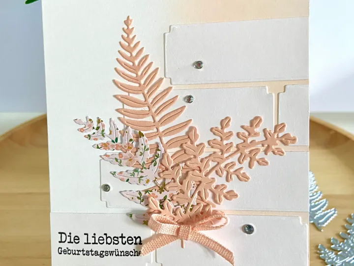 Eine gebastelte Geburtstagskarte mit gestanzten Farnblätter in Sand- und Pastellgrüntönen, die über eine weiße Grundfläche mit beige Akzenten angebracht sind. Ein feiner Seidenfaden in Sandfarbe ist als Schleife am unteren Ende der Blätter befestigt. Kleinste Kristallsteine dienen als Verzierung an den Rändern der Kartenfelder. Der Text „Die liebsten Geburtswünsche“ ist im unteren Bereich mit schwarzer Schrift aufgedruckt. Im Hintergrund sind grüne Pflanzenblätter und weitere gestanzte