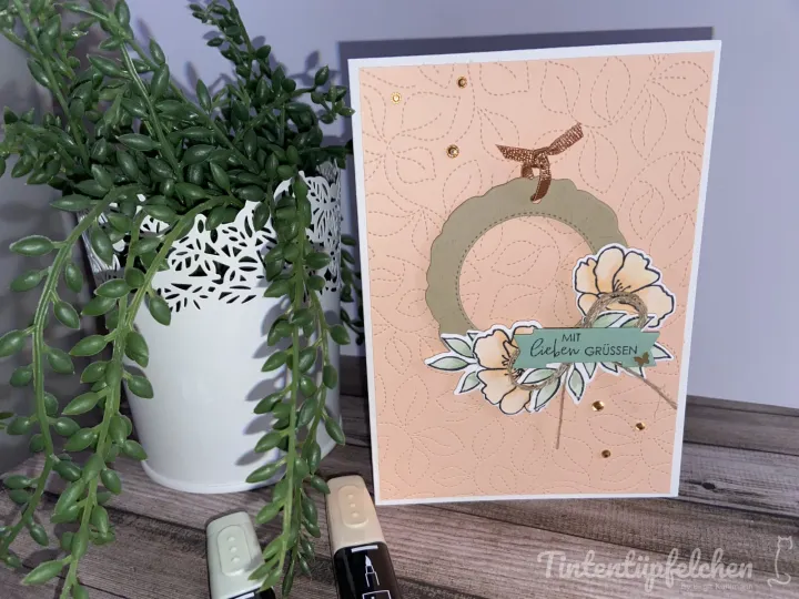 Eine selbstgebastelte Grußkarte mit gestempelten Blumen in Pastellfarben und einem grünen Kreis auf helloranger Basis, verziert mit kleinen goldenen Punkten und einer Beschriftung „Mit lieben Grüßen“. Links daneben steht eine Pflanze in einem weißen Topf mit Ausschnittsmuster.