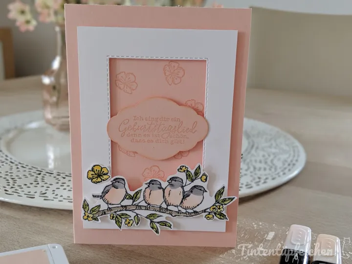 Eine gebastelte Geburtstagskarte mit rosa und weißem Papier, gestochenen Vögeln auf einem Ast und gelben Blüten. Im Zentrum befindet sich ein ovaler Textblock mit der Aufschrift „Ich sing dir ein Geburtstagsslied, denn es ist Gechen, dass es dich gibt!“. Der Hintergrund zeigt eine Tischplatte mit Tintenflaschen und einem Glas mit Blumen.