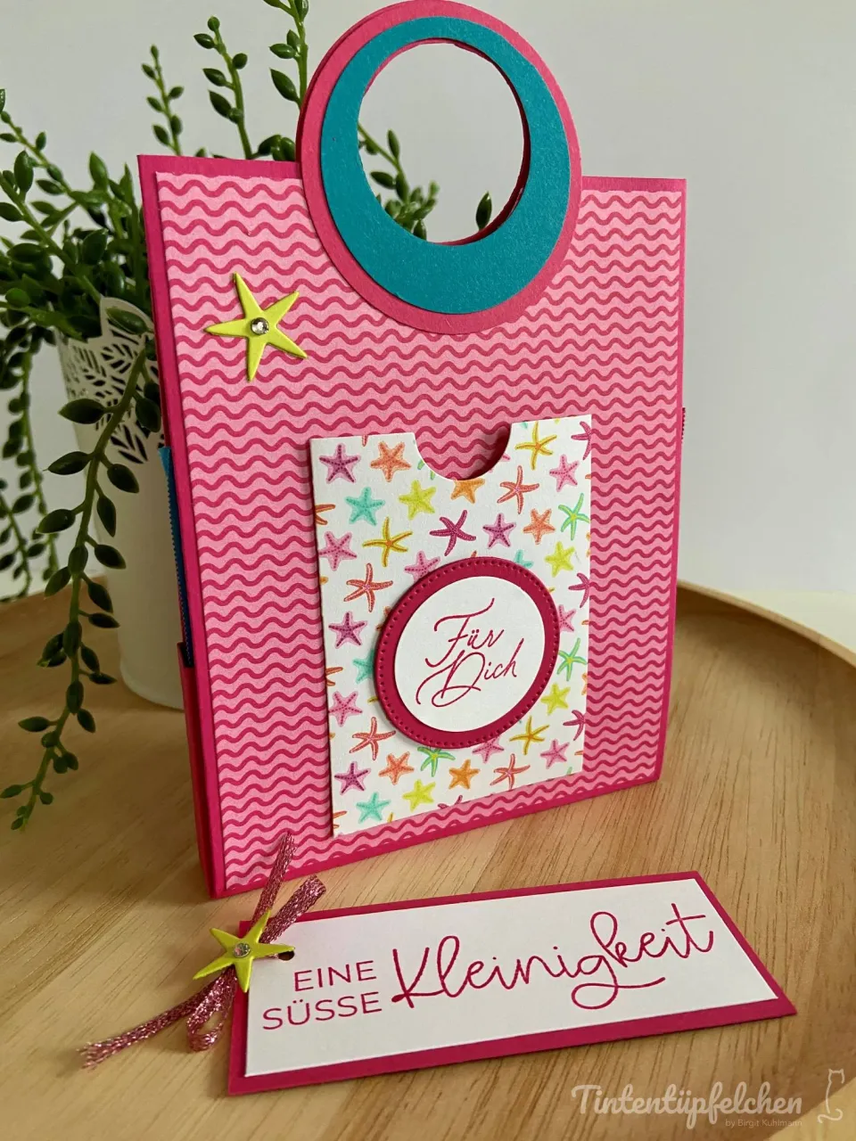 Ein gebasteltes Geschenkpaket aus rosa Papier mit Wellenmustern und einem blauen Ring als Griff, dekoriert mit bunten Sternen und einer Karte „Für Dich“. Vor dem Paket liegt ein Beschriftungssticker mit der Aufschrift „Eine SÜSSE Kleinigkeit“ in roter Schrift.
