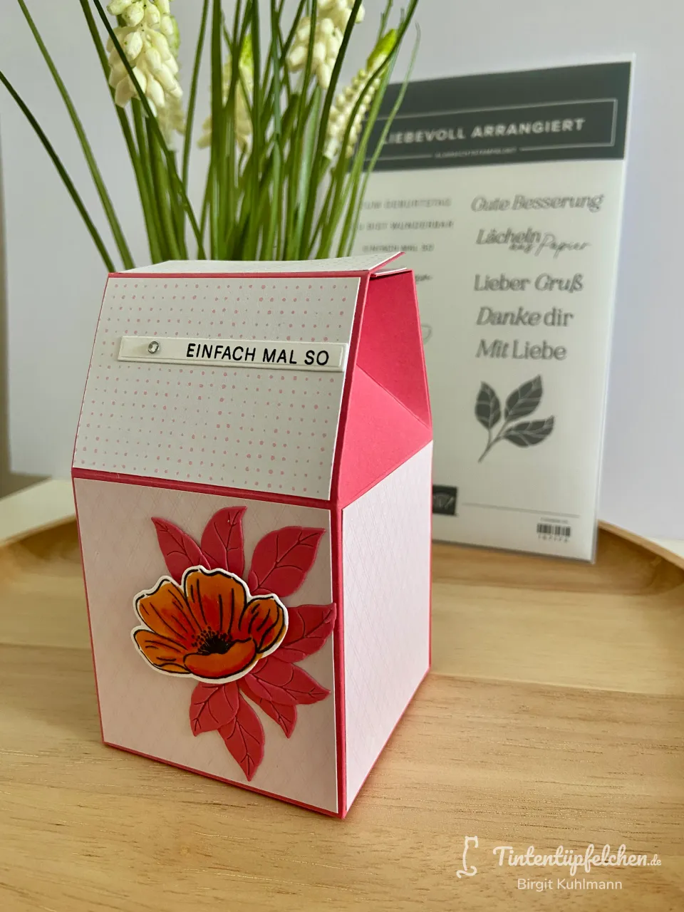 Ein gestempelter Kasten in Rosa mit weißem Punktmuster und einer orangefarbenen Blume als Aufkleber. Im Hintergrund steht ein Stempelset mit deutschen Grußwörtern vor einem Vasenarrangement mit grünen Pflanzenstängeln.