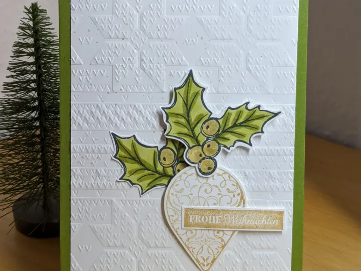 Eine grüne Klappkarte mit einer weißen Prägekarte, die mit einem Muster aus kleinen Dreiecken versehen ist. Darauf sind gestempelte und koloriere Stechpalmenblätter mit Beeren sowie ein goldfarbener Anhänger mit der Aufschrift "Frohe Weihnachten" angebracht.